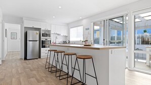 Ett modernt kök med vita skåp, ett kylskåp i rostfritt stål, en mikrovågsugn och en inbyggd ugn. Det finns fyra barstolar vid köksön och ett stort fönster med persienner.