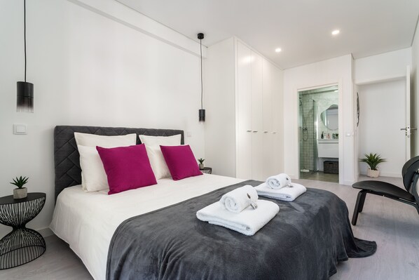 Habitación de hotel moderna con una cama grande, una silla, una mesita y un baño visible a través de una puerta abierta.