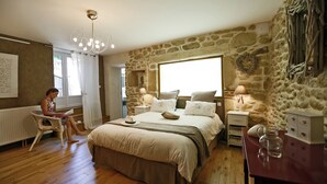 Ein Schlafzimmer mit Steinwand, einem großen Bett mit vielen Kissen, einem Nachttisch mit Lampe und einem Sessel, in dem eine Person liest.