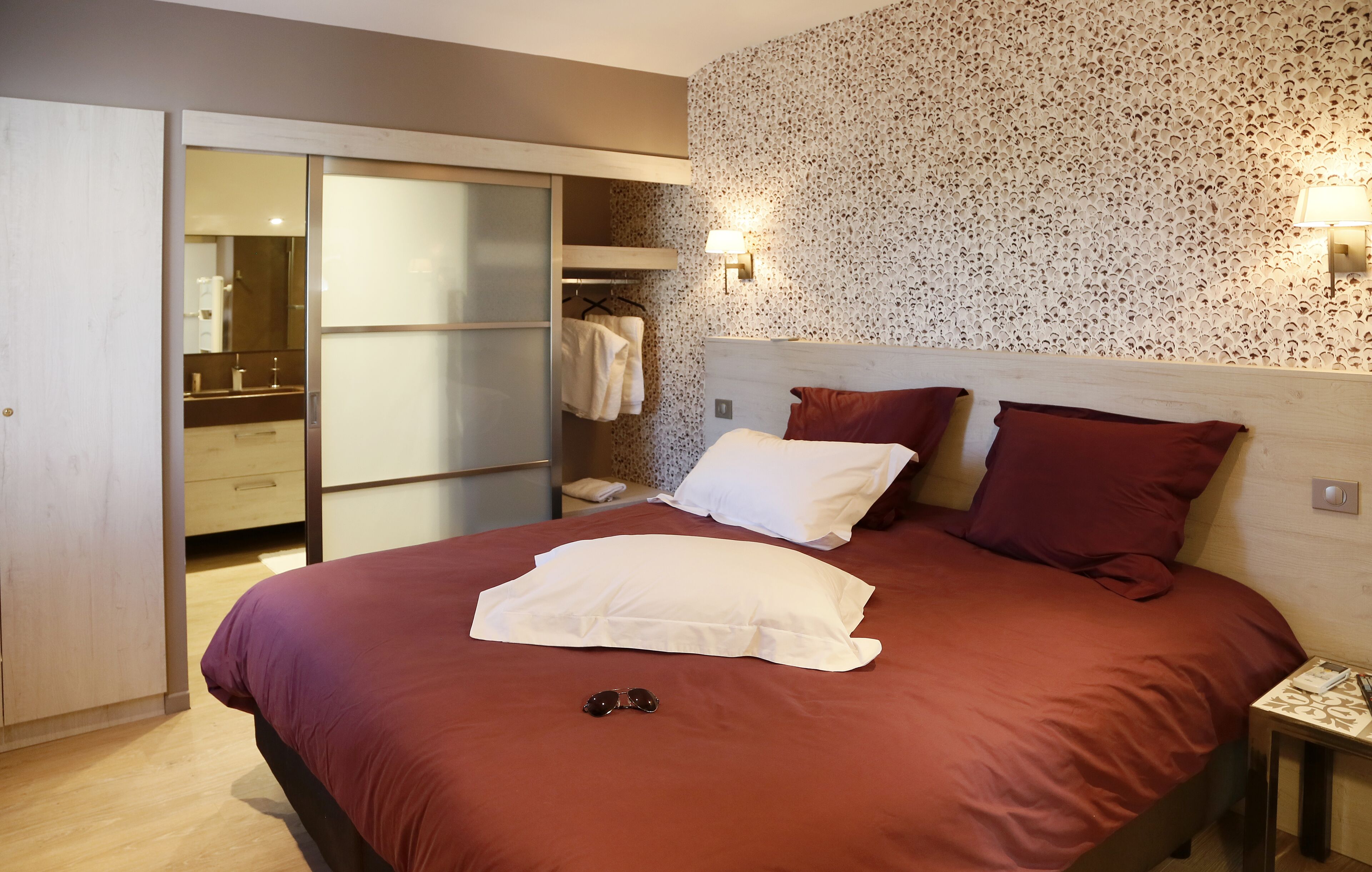 Une chambre à coucher avec un grand lit, deux oreillers, une table de chevet, un miroir, une armoire et une lampe fixée au mur.