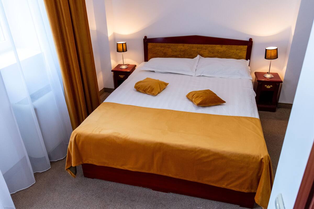 Una habitación de hotel con una cama grande, dos mesitas de noche con lámparas y una ventana con cortinas.