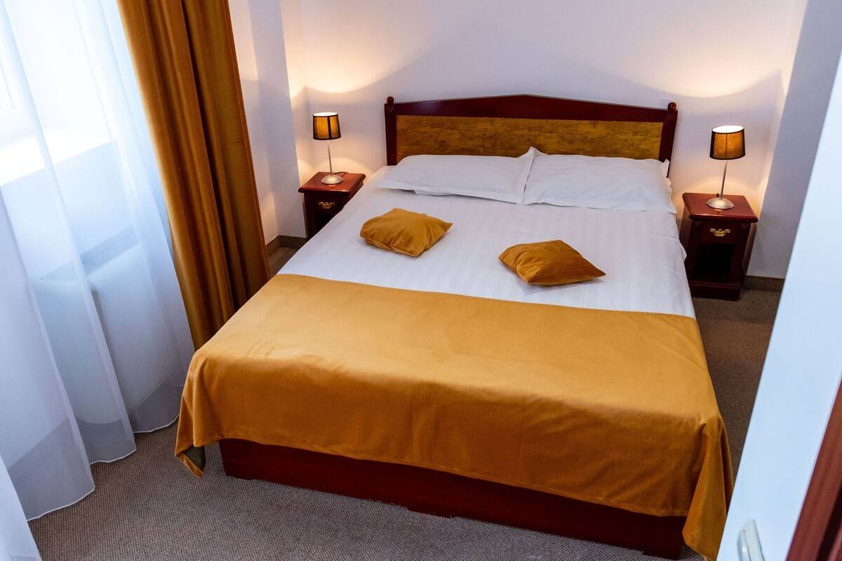 Habitación de hotel con una cama, dos mesitas de noche, una ventana con cortinas y suelo alfombrado.