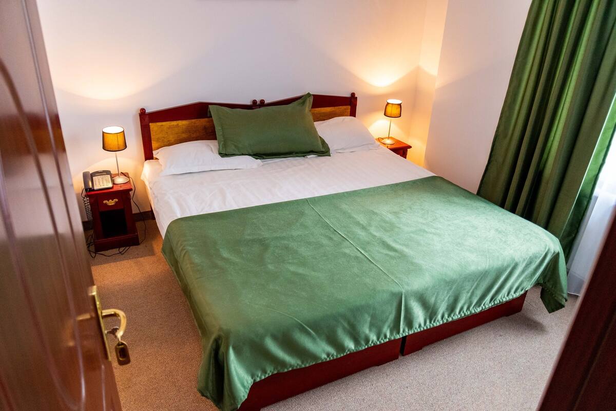Habitación de hotel con cama, lámparas de noche, un teléfono y una cortina verde.
