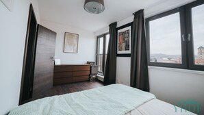 A bedroom with a bed, a dresser, a map on the wall, and a window with a view of buildings. - Enjoy panoramic view of The Little Carpathian (Pezinok)