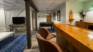 Une pièce avec un bar en bois, des fauteuils en cuir marron, une table de baby-foot, un téléviseur et un escalier.