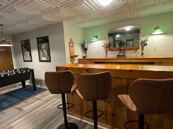 A bar area with wooden stools, a foosball table, and wall-mounted green lamps. - Beautiful Pines Tucked in the trees, minutes to Cooperstown & Dreams Park (Cooperstown)