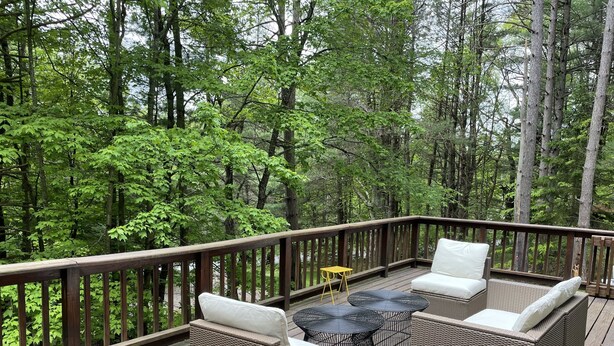 Une terrasse en bois, agrémentée de meubles en osier et d’une petite table, entourée d’une forêt dense.