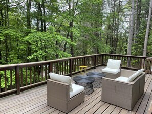 A wooden deck with wicker furniture and a small table, surrounded by dense forest. - Beautiful Pines Tucked in the trees, minutes to Cooperstown & Dreams Park (Cooperstown)