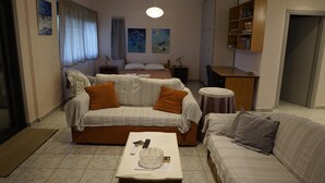 A living room with a sofa, a smaller couch, a coffee table with a glass bowl and remote controls, a bed with pillows, and a bookshelf. - Lovely Modern Studio in Romanos Patras, Greece (Patras)
