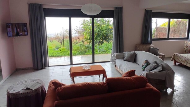A spacious living room with a large sliding glass door leading to a garden, a sofa, a small table, and a television mounted on the wall. - Lovely Modern Studio in Romanos Patras, Greece (Patras)
