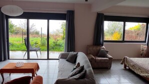 A living room with a sofa, armchair, and a small table. There are large windows with curtains, and a view of a garden. - Lovely Modern Studio in Romanos Patras, Greece (Patras)
