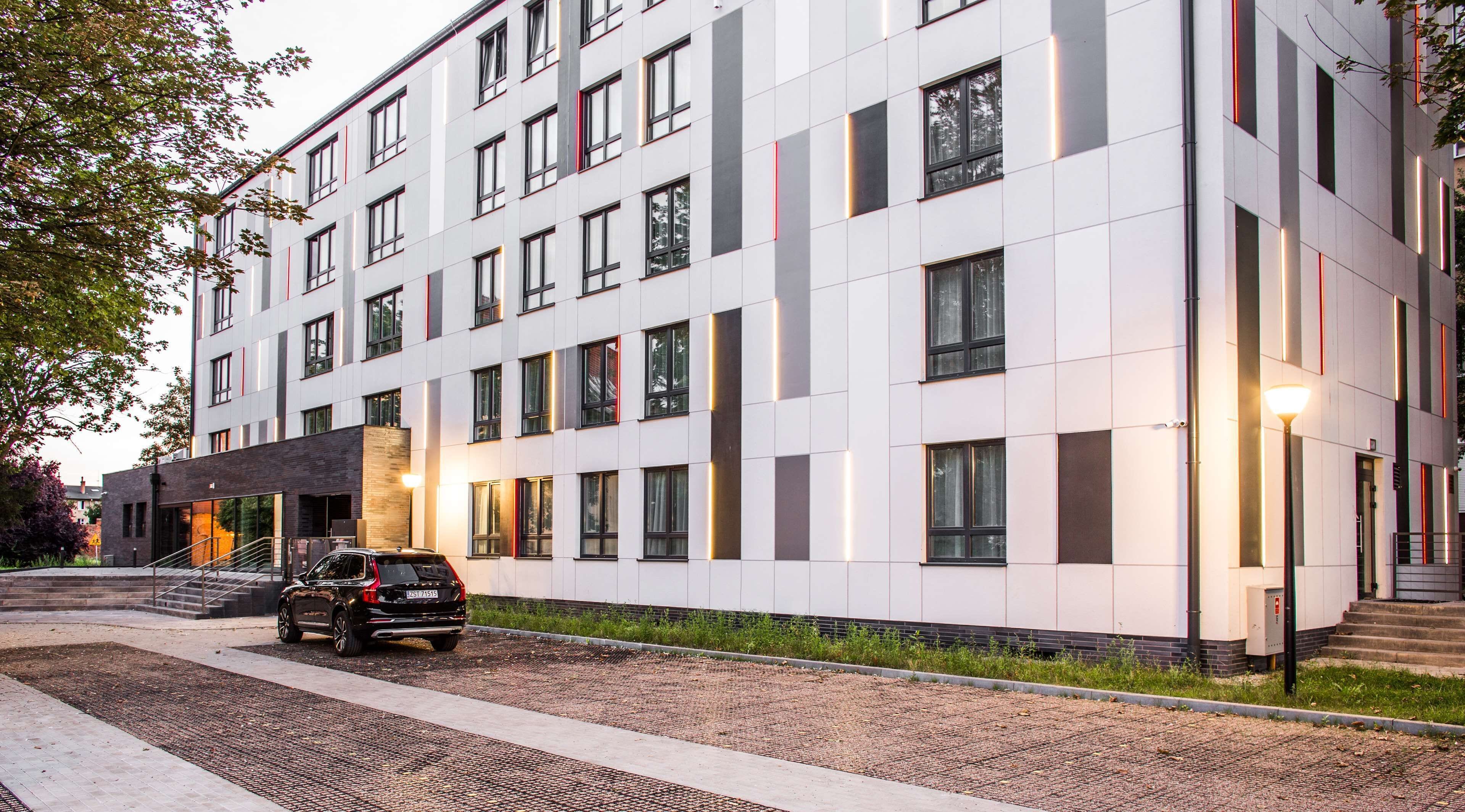 A modern multi-story building with a clean, white facade, large windows, and a car parked in front.