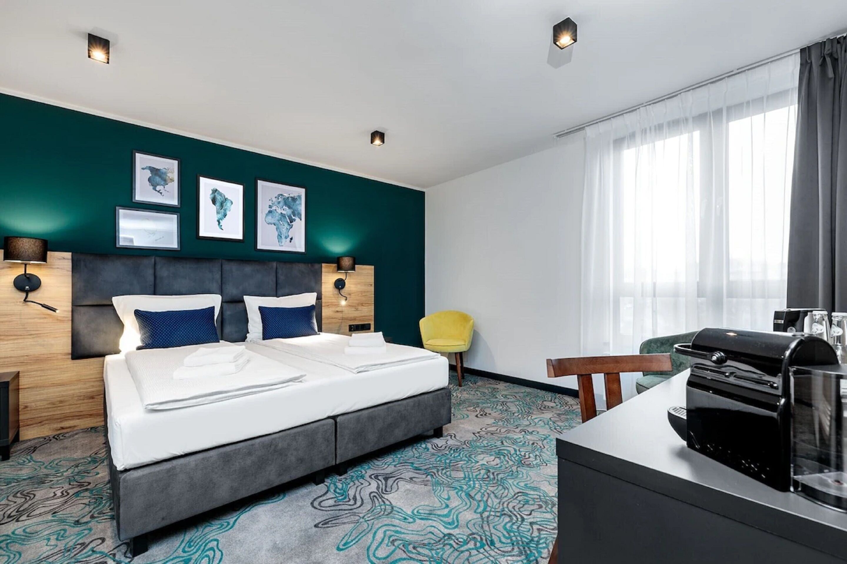 Una habitación de hotel moderna con una cama grande, un escritorio con cafetera, una silla y obras de arte en las paredes.