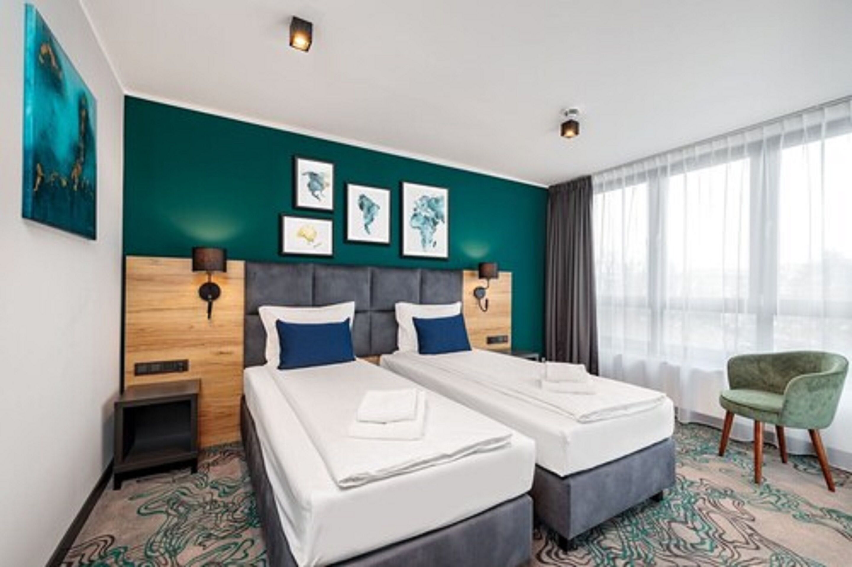 A hotel room with two beds, a green accent wall, artwork, a green chair, and a view of the outdoors.