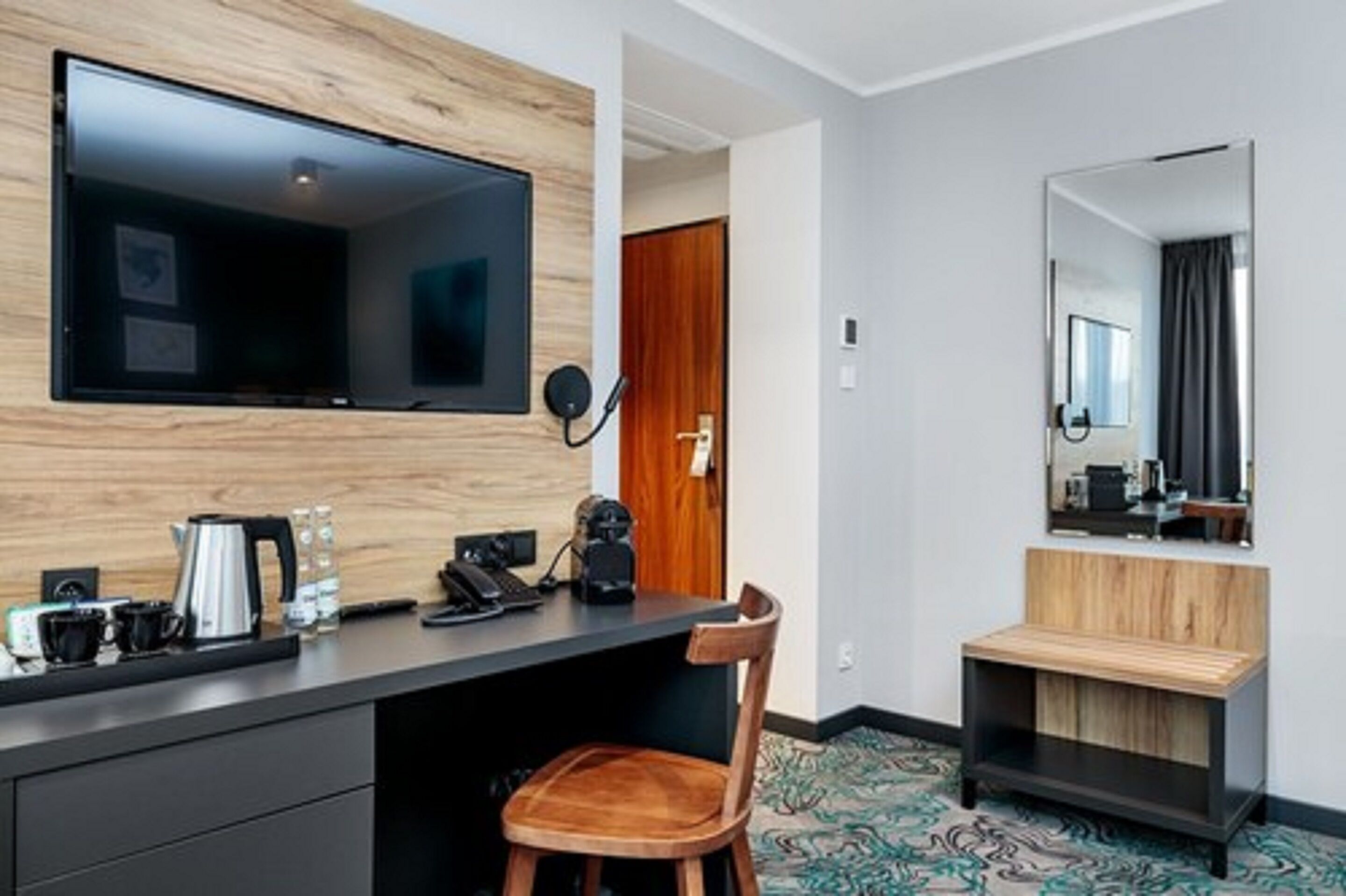Una habitación de hotel moderna con televisión de pantalla plana en una pared de madera, un escritorio con cafetera, una silla y un pequeño banco de madera.