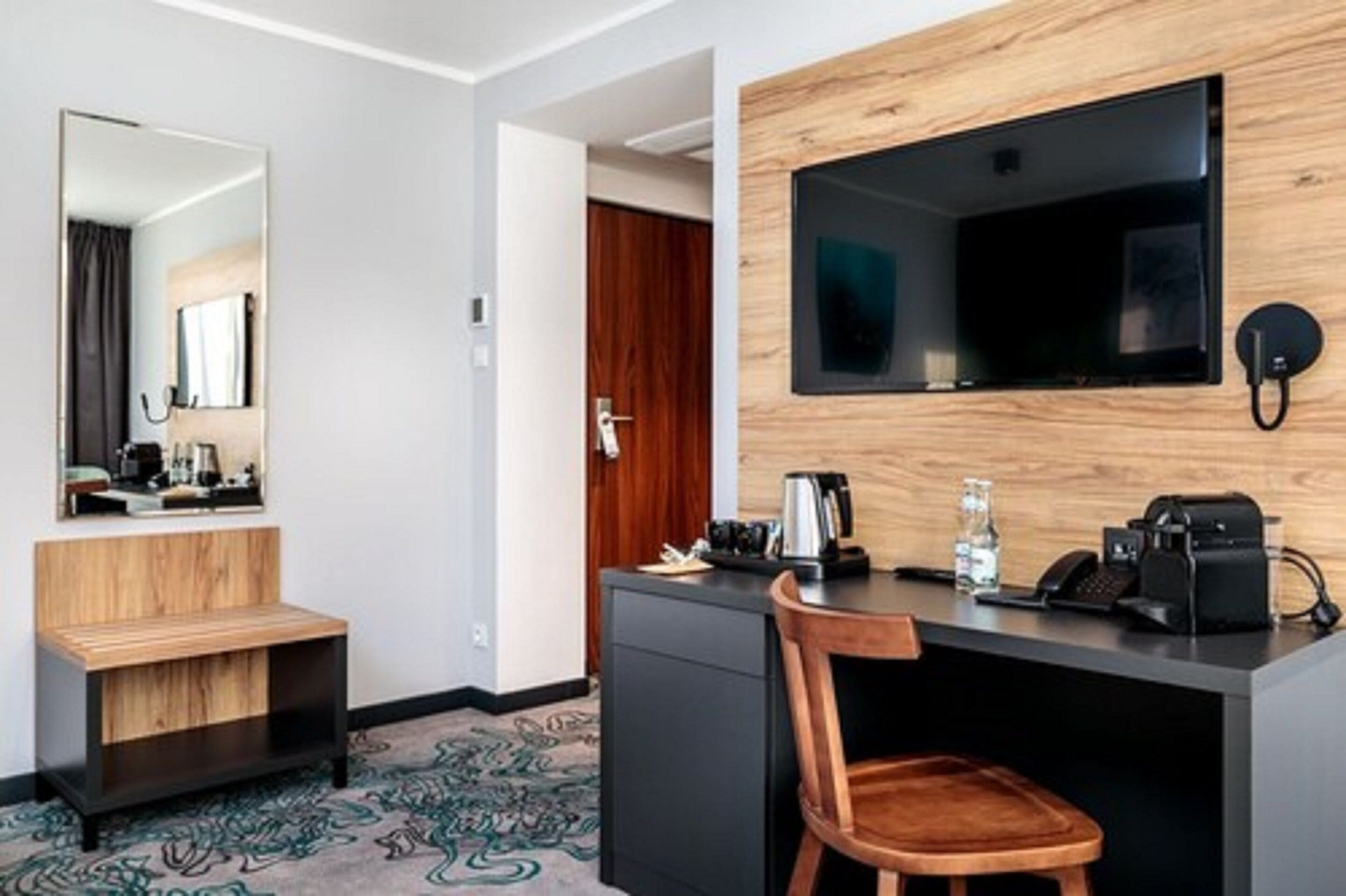 Hotelkamer met een houten bankje, een bureau met telefoon, een koffiezetapparaat en een flatscreen televisie bevestigd aan een houten wand.