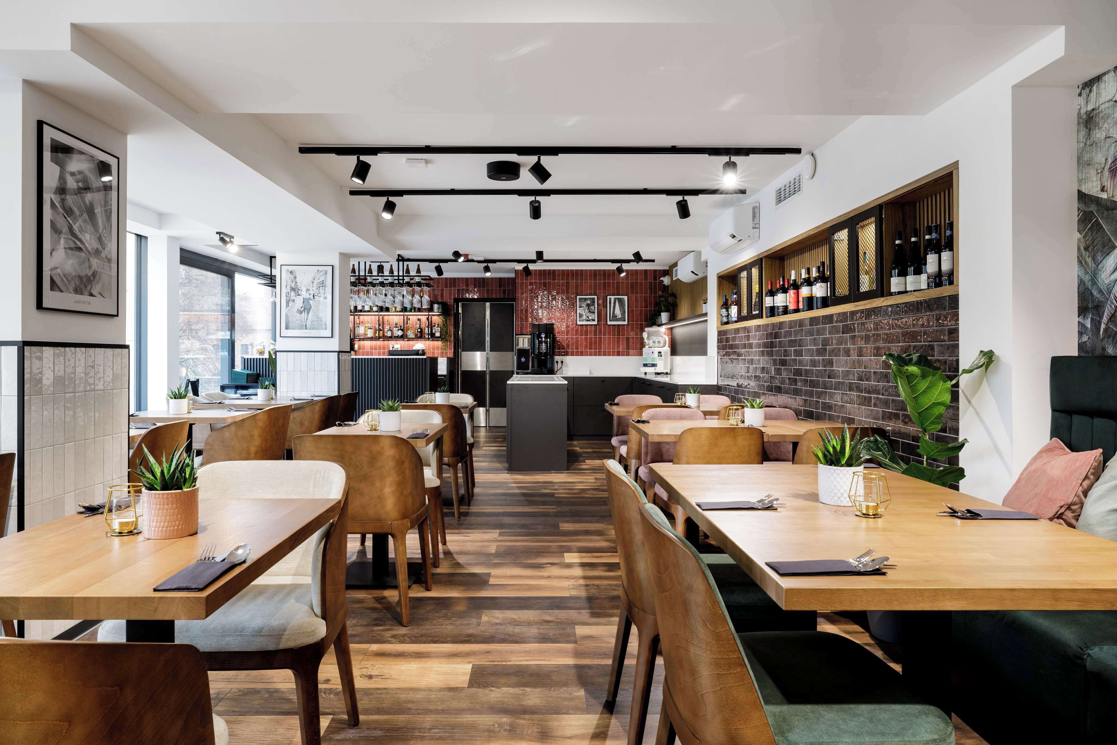A modern restaurant with wooden tables and chairs, a bar area with wine bottles, and a kitchen in the background.