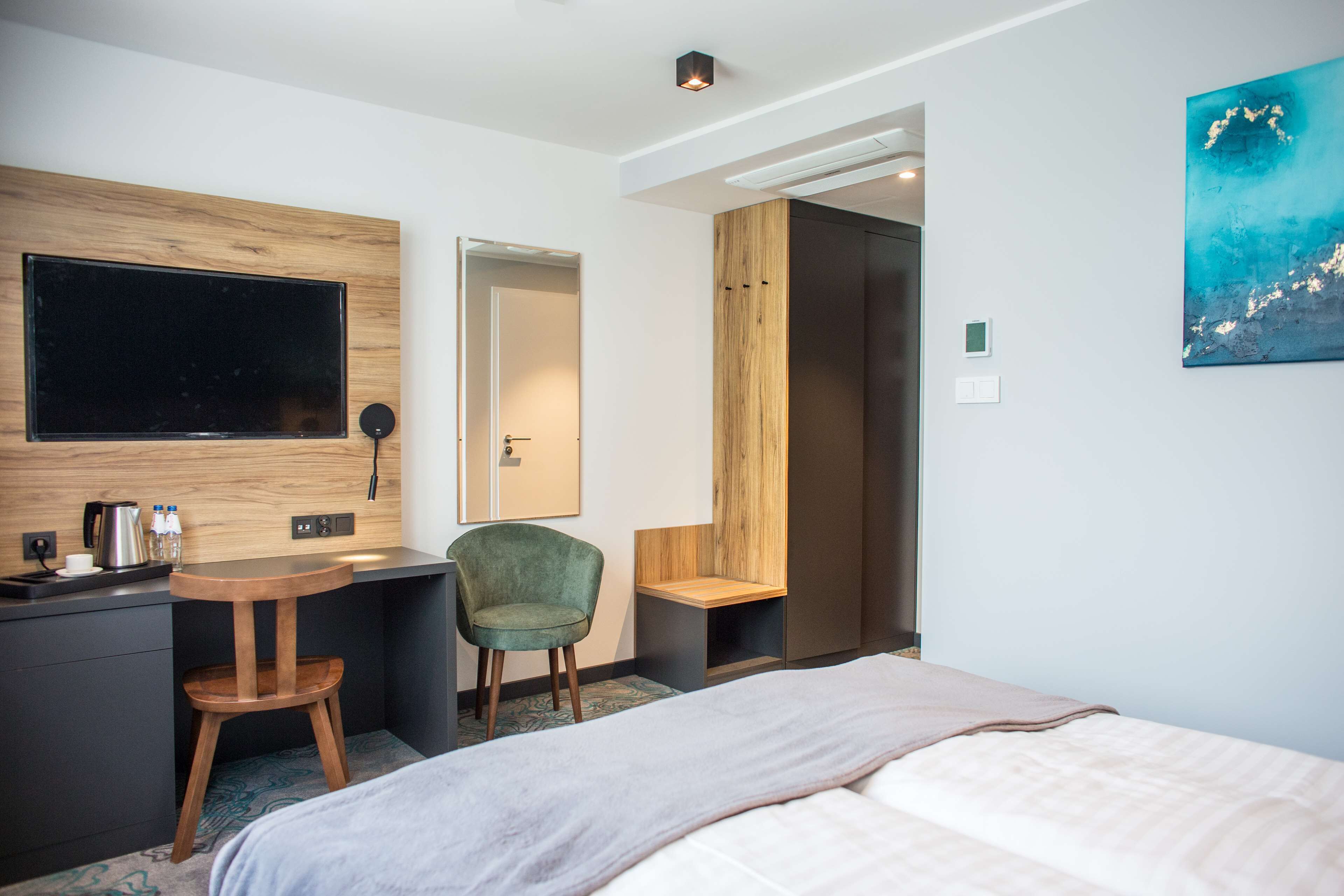 Hotellihuone, jossa on sänky, työpöytä tuolilla, taulutelevisio, kahvinkeitin ja taulu seinällä.