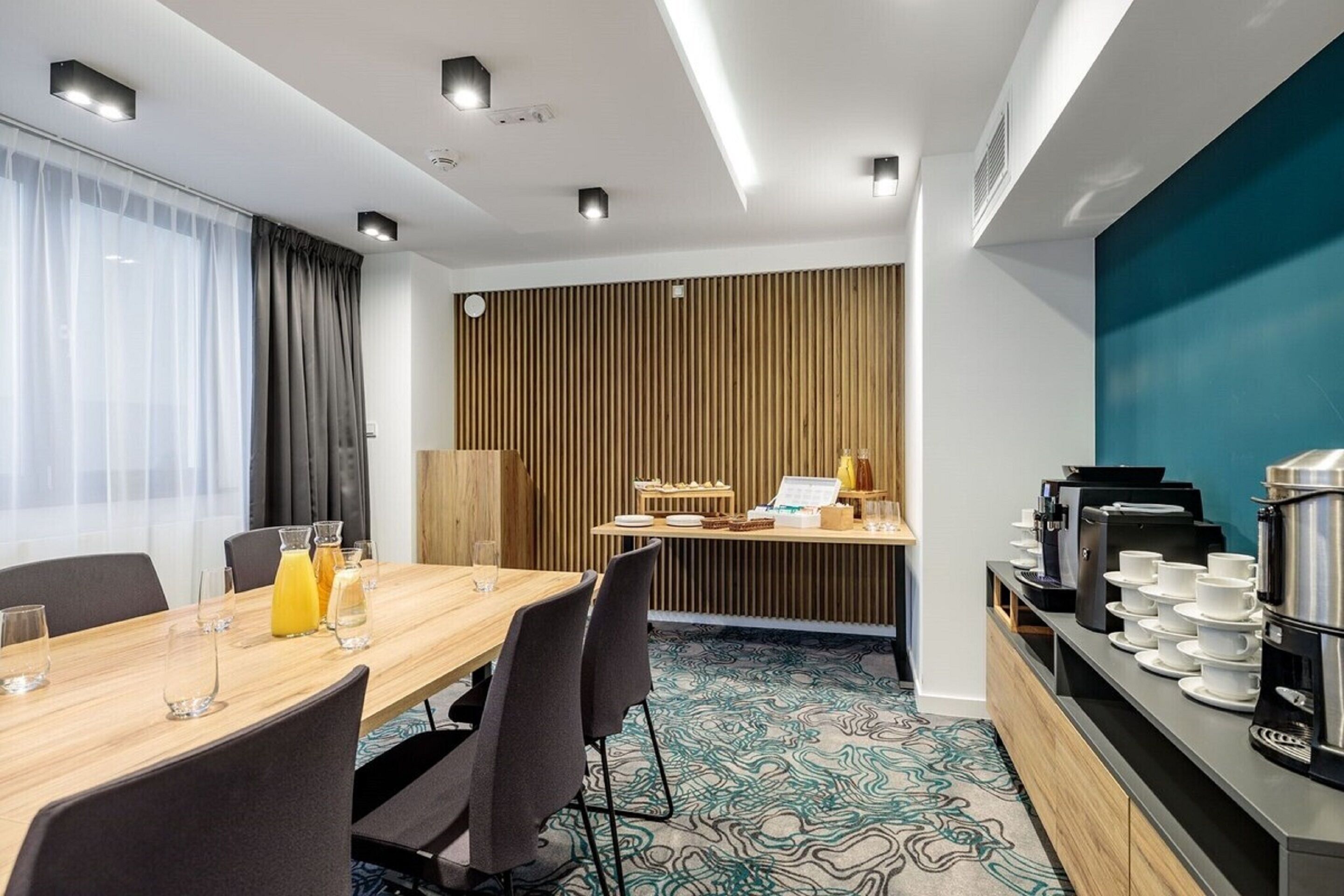 A hotel room with a wooden dining table, chairs, a coffee station, and a view of the city.