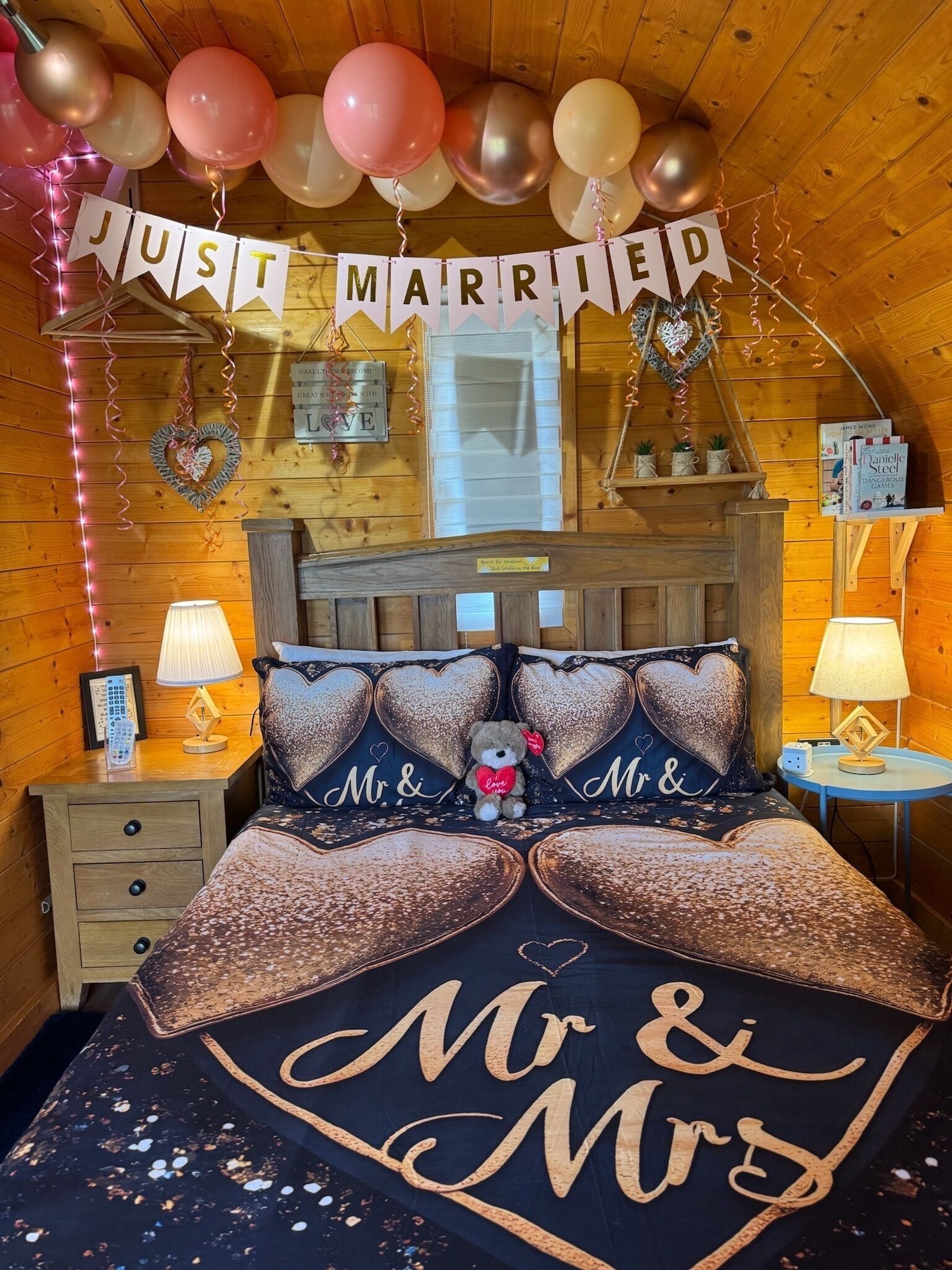 A cozy, wooden-themed bedroom with a 'Just Married' banner, heart-shaped pillows, and a bedspread with 'Mr & Mrs' lettering.