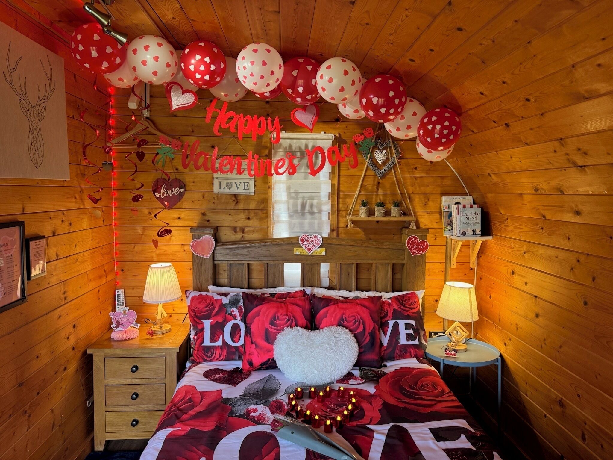 A cozy bedroom with a wooden ceiling, a bed with red and white floral bedding, heart-shaped pillows, and a 'Happy Valentine's Day' sign.