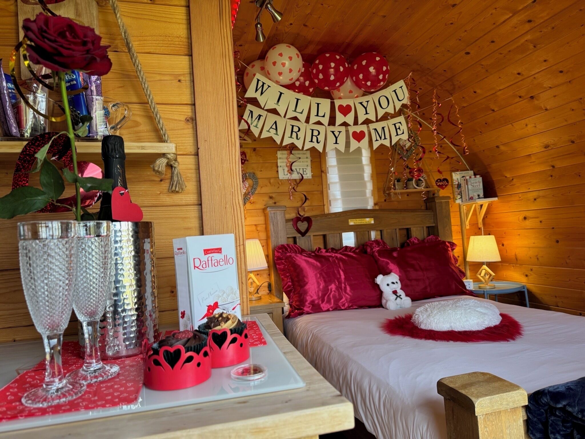 Una habitación acogedora con una cama, una mesa con pastel y vasos, y un cartel que dice '¿Te casarías conmigo?'
