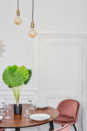 Un comedor con una mesa redonda de madera dispuesta para cuatro personas, una planta en maceta y una silla rosa.