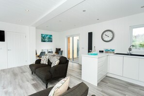 A modern living room with a sofa, a dining area with chairs, a kitchen with a sink and a clock, and a sliding glass door leading to an outdoor area.