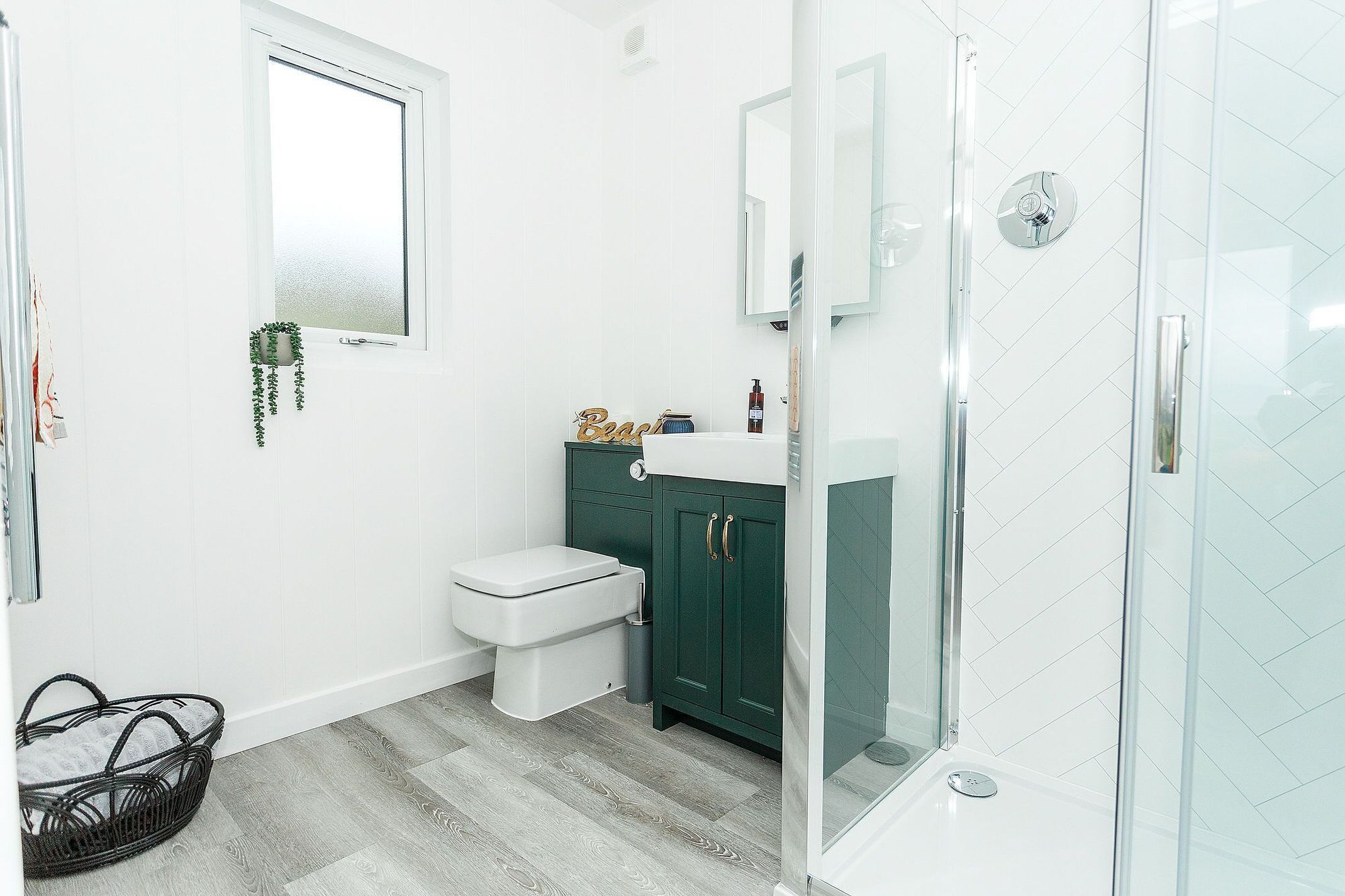 Een badkamer met een wit toilet, groene kastjes, een douchecabine met glazen wanden en een vloer van witte tegels.