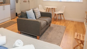 A modern living room with a sofa, two chairs, a small table, and a kitchen area with cabinets and a refrigerator. - The Mew (Dartmouth)