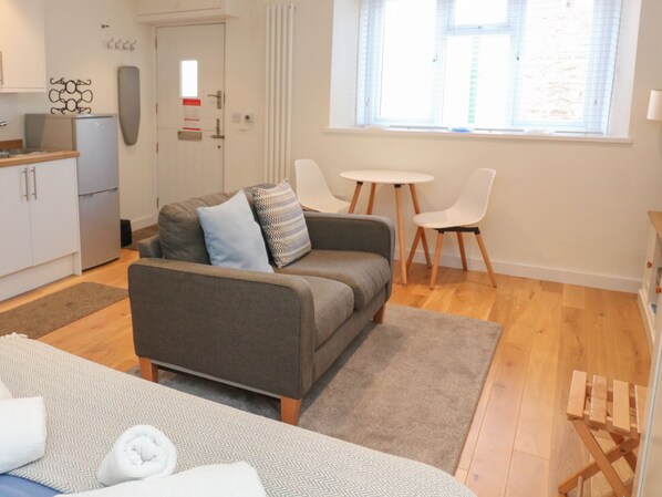 A modern living room with a sofa, two chairs, a small table, and a kitchen area with cabinets and a refrigerator. - The Mew (Dartmouth)
