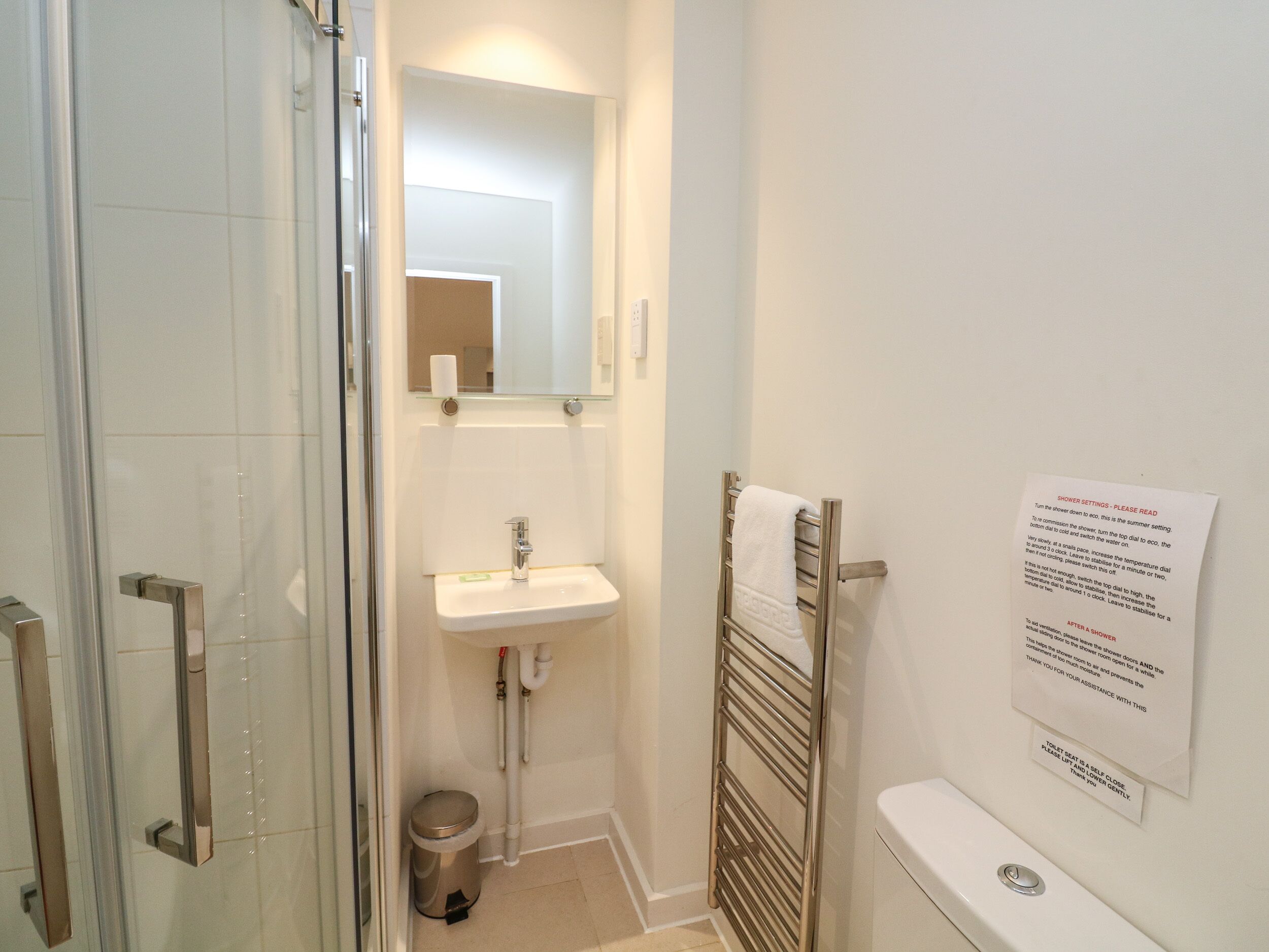 Een badkamer met een douchecabine van glas, een wastafel met spiegel erboven, een handdoekhouder en een bericht op de muur.