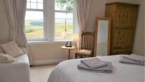 A bedroom with two beds, a wooden wardrobe, a chair, a small table with a lamp, and a view of the countryside through the window.