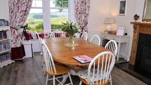 A dining area with a wooden table, white chairs, and a view of the countryside through large windows.