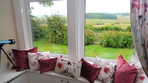 A room with a window seat adorned with patterned cushions, a telescope, and a view of a green landscape.