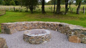 A stone fire pit with a metal grill, surrounded by gravel and large rocks, set in a grassy outdoor area with trees and a fence in the background.