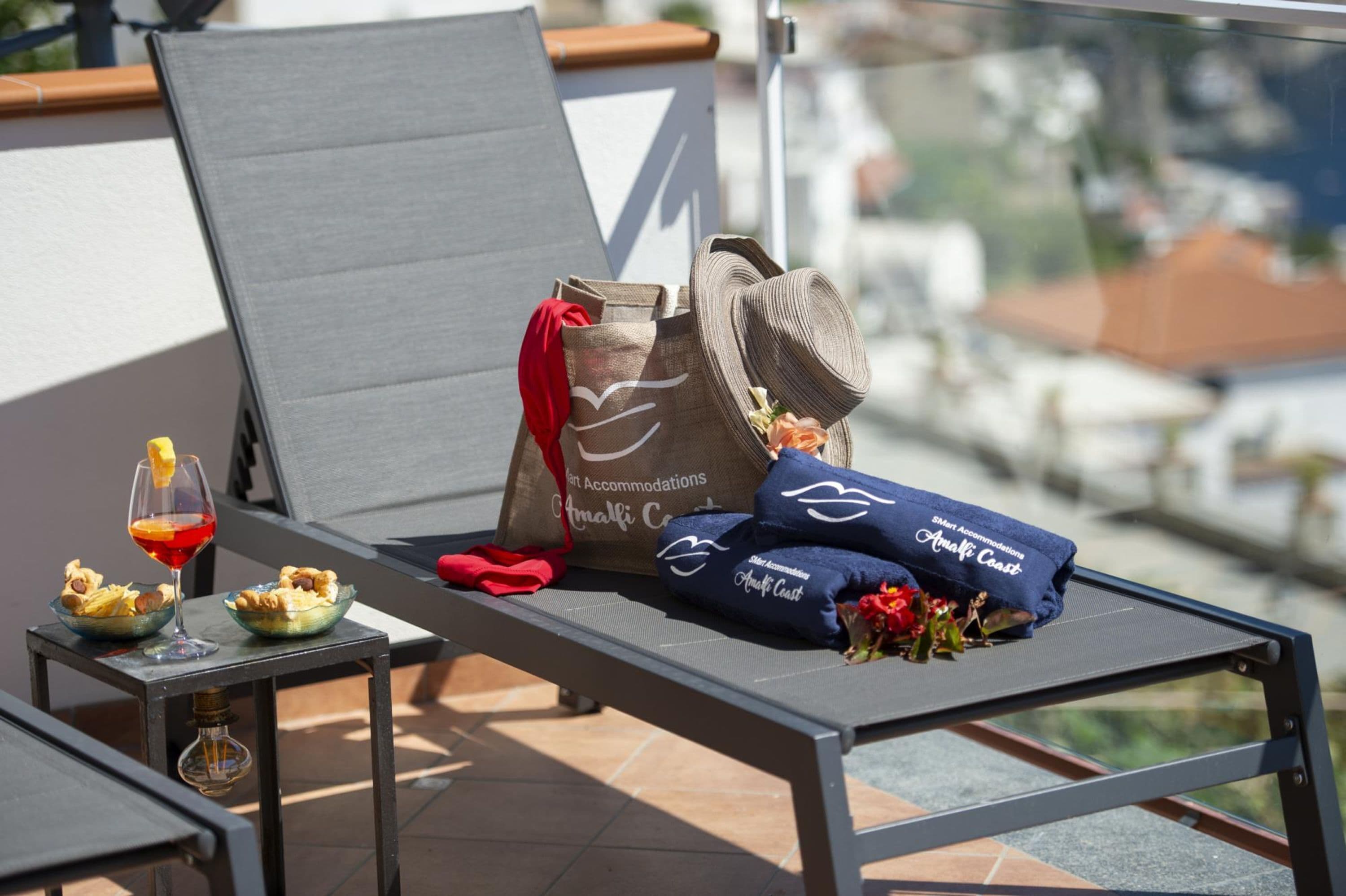 A sun lounger with a view of the city, a glass of red wine, snacks, a bag, and towels on a balcony.