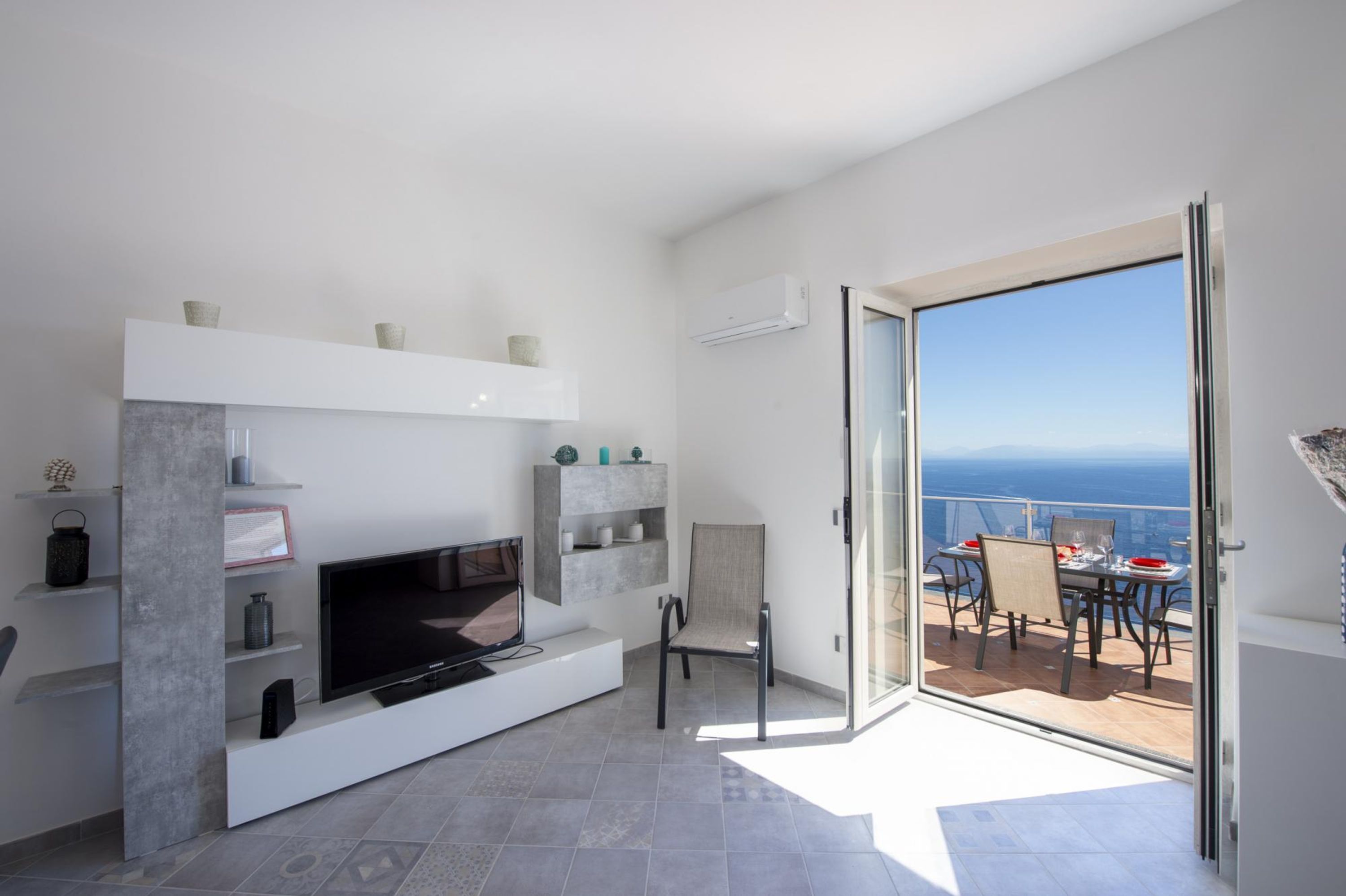 Een moderne woonkamer met een flatscreen televisie, inbouwkasten en een glazen deur naar een balkon met uitzicht op zee.