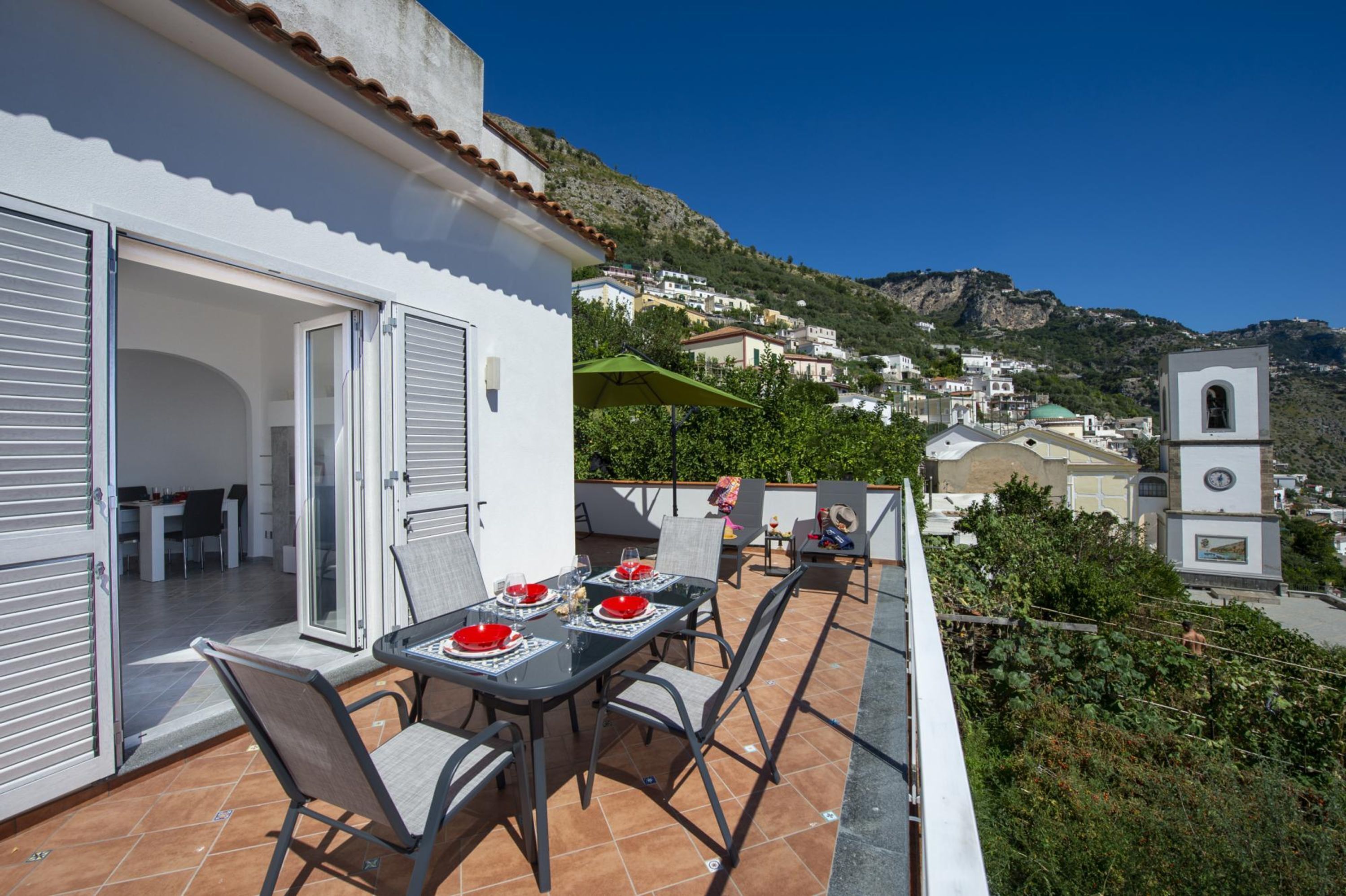 Un balcón con una mesa lista para comer, una puerta corrediza de vidrio que da a una habitación interior, y vista de un pueblo en la ladera con edificios y una iglesia.