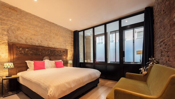 Une chambre à coucher moderne avec un grand lit, une tête de lit en bois, une table de chevet avec une lampe, un fauteuil jaune et une grande fenêtre donnant sur la rue.