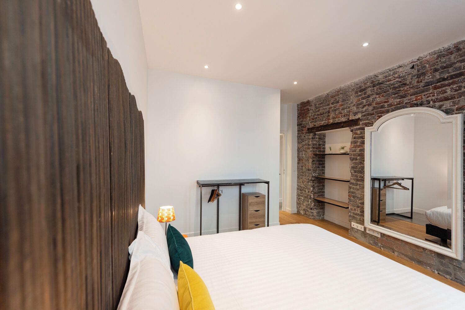 Quarto com uma cama grande, cabeceira de madeira, escrivaninha com gavetas, espelho e parede de tijolos.