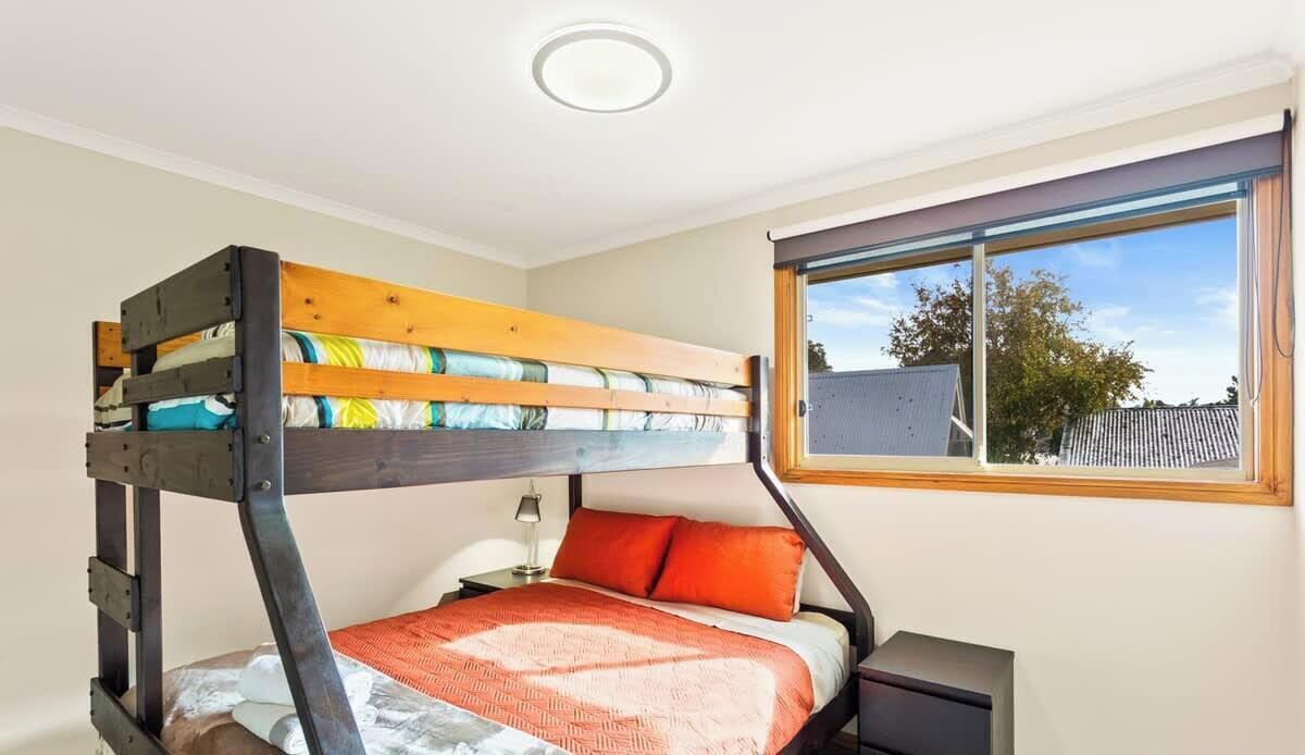 A bunk bed with a wooden frame, a bedside table, a lamp, and a window with a view of the outside.