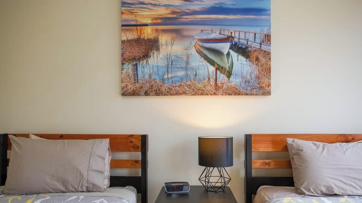 Quarto de hotel com duas camas, uma mesa de cabeceira com luminária e um quadro de um barco em um cais.