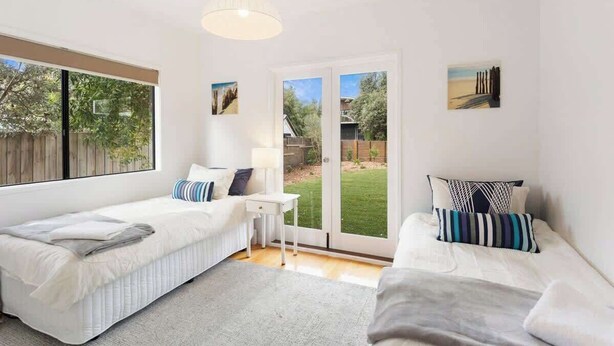 A room with two beds, a small table, a lamp, and a sliding glass door to the outside. - Serene, Modern Beach Escape 3 Bed 2 Bath (Cape Woolamai)