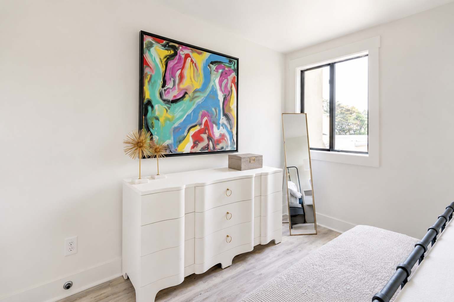 Un dormitorio con un aparador blanco, un cuadro abstracto grande, un espejo de cuerpo entero y una ventana.