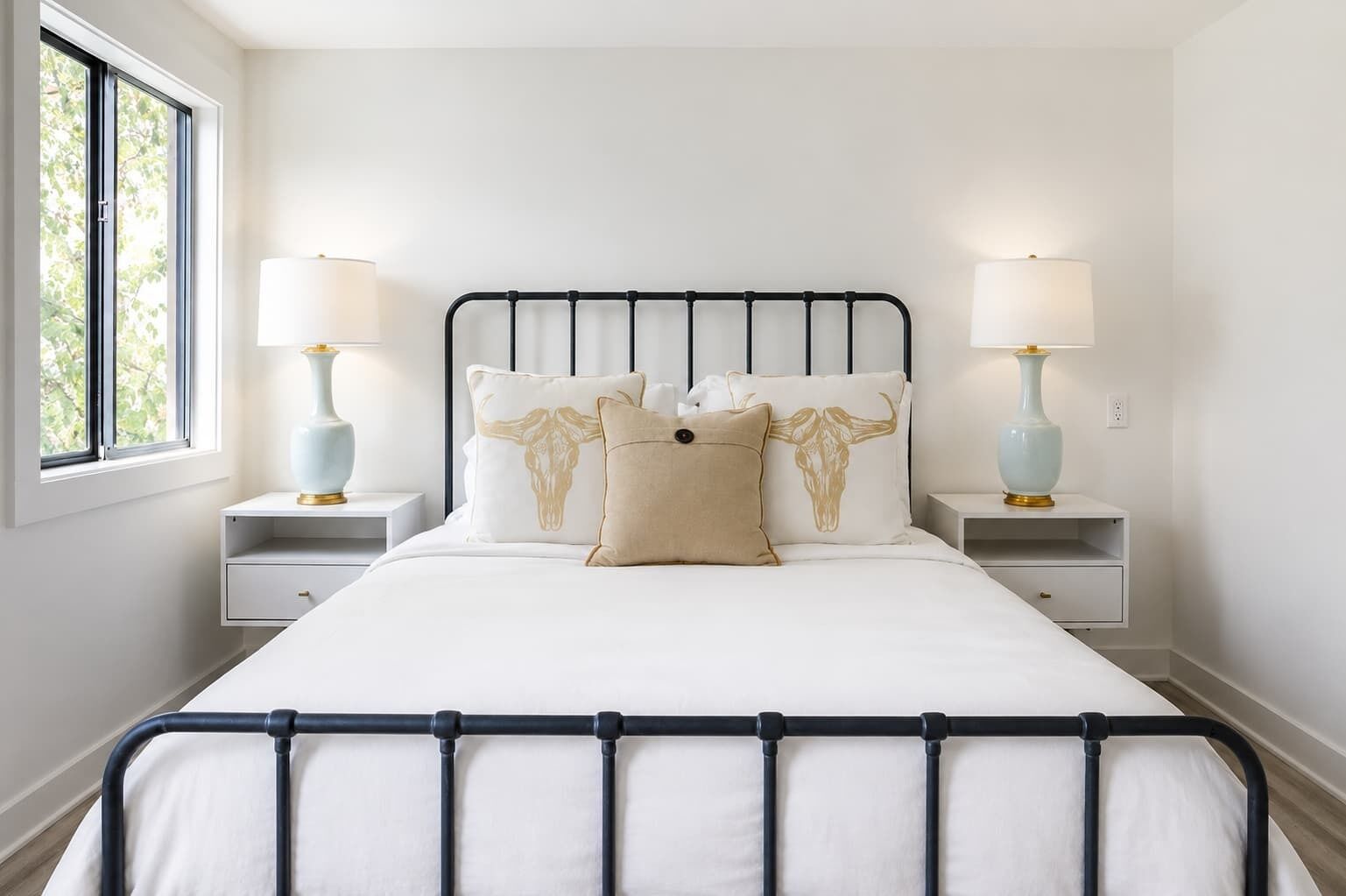 A neatly made bed with a metal frame, two bedside tables with lamps, and a window with a view of trees.