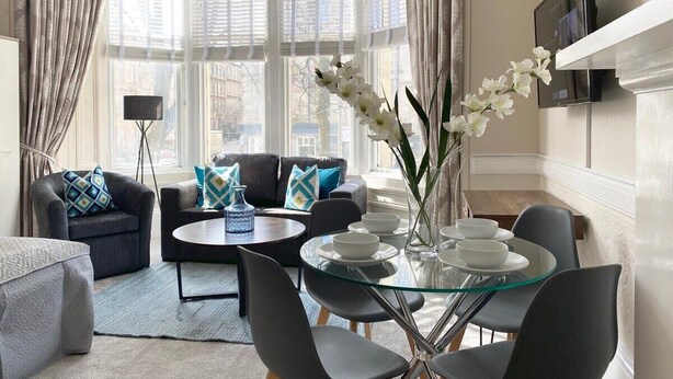 A modern living room with a glass dining table, black chairs, a sofa with patterned cushions, and a large window with blinds.
