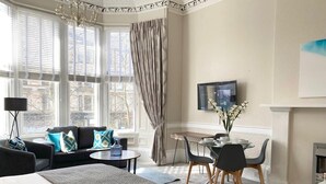 A hotel room with a large bed, a sofa, a dining table with chairs, a TV, and a fireplace.