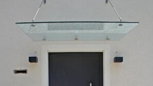 Entrada de un edificio moderno con una puerta oscura, un panel de vidrio arriba y una barandilla blanca a ambos lados.
