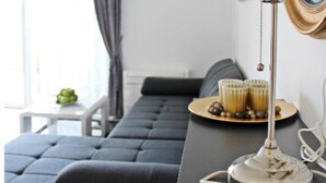 Una sala de estar moderna con un sofá oscuro, una mesa redonda con objetos decorativos, una lámpara blanca y un espejo grande.